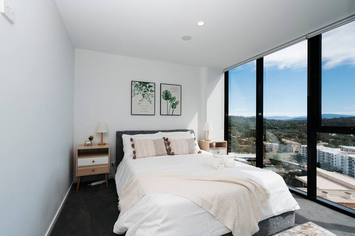 The spacious master bedroom offers a serene retreat with a king-sized bed dressed in luxurious linens. The large window frames a breathtaking cityscape, providing a stunning backdrop for your stay. Perfect for relaxation or enjoying the urban view.