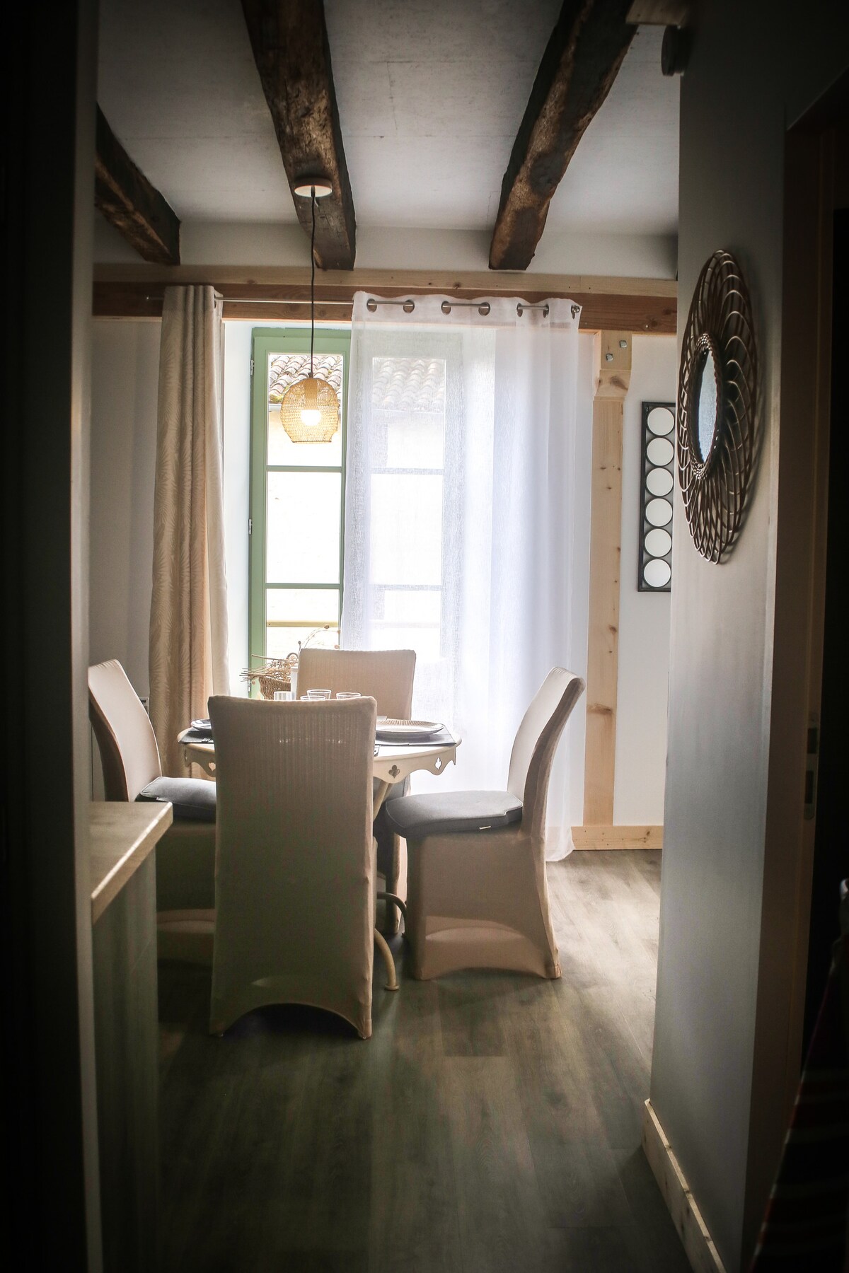 A dining area is featured with a round table surrounded by four chairs. Natural light enters through a large window, adorned with sheer curtains. Exposed wooden beams add character to the ceiling, enhancing the space's overall charm.