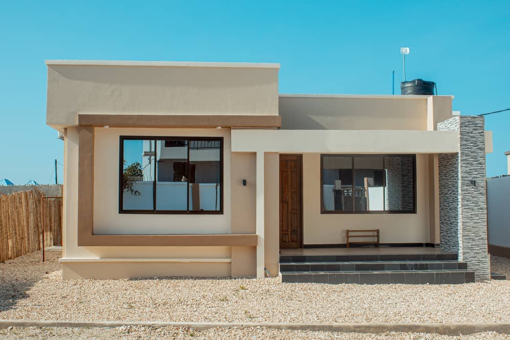 A modern single-story home features a clean, minimalist design with a smooth beige exterior. A large window allows natural light to fill the interior, while a stone accent complements the entryway. A small bench is positioned on the front porch, enhancing the welcoming vibe.
