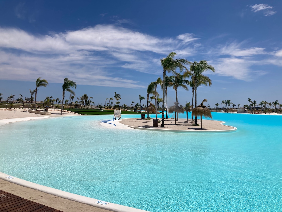 A large artificial lagoon with clear turquoise water spans across the image, bordered by sandy shores. Palm trees provide scattered shade around the lagoon’s edges. The bright, sunny sky features a few clouds, enhancing the serene outdoor setting.