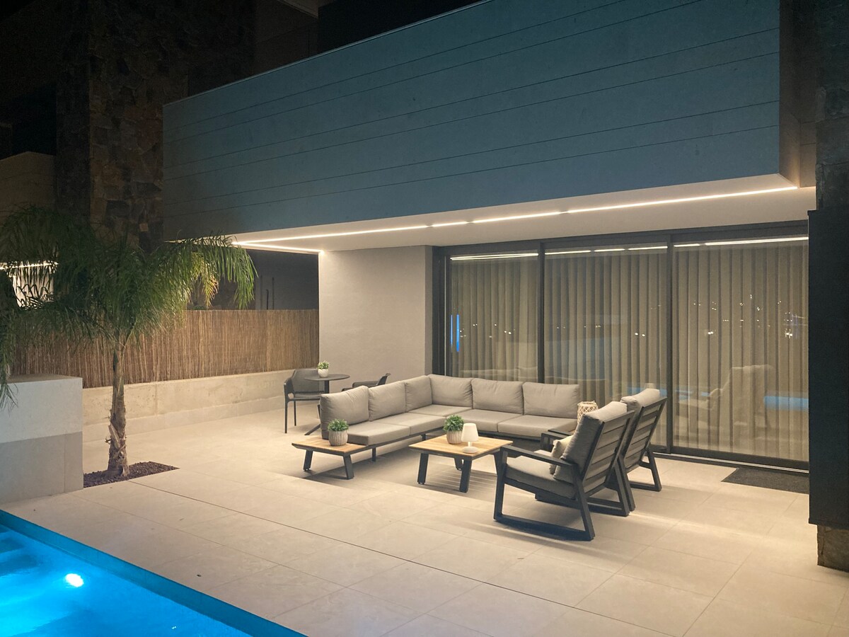 An outdoor seating area features a modern sectional sofa and several chairs, arranged around small decorative tables. The space is illuminated by soft lighting, with a serene pool visible nearby, and large glass doors providing a view into the villa's interior.