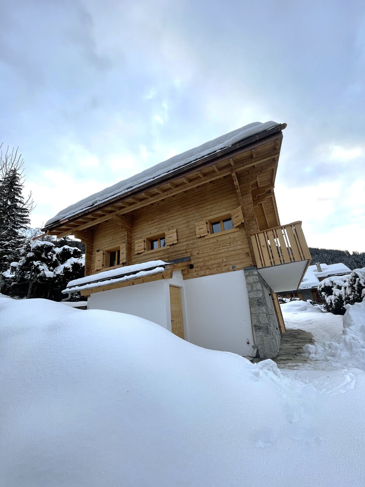 A wooden chalet is seen against a cloudy sky, set amidst a snowy landscape. The structure features large windows and a small balcony, blending rustic charm with its natural surroundings. Snow blankets the ground, creating a serene winter scene.