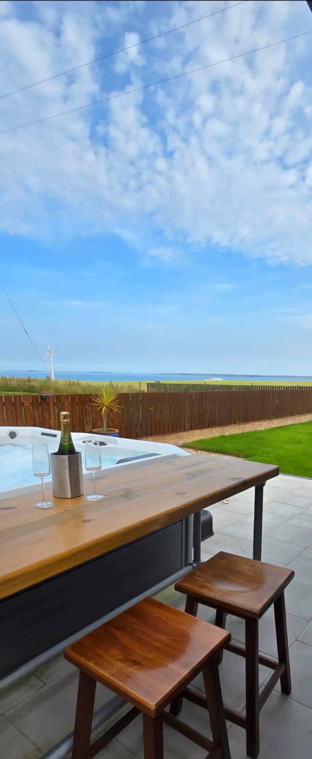 An outdoor setting features a wooden bar table accompanied by two stools, overlooking a scenic view of the sea and sky. A bottle of champagne and two glasses rest on the table, enhancing the inviting space. The grassy area in the foreground leads towards a fenced backdrop.