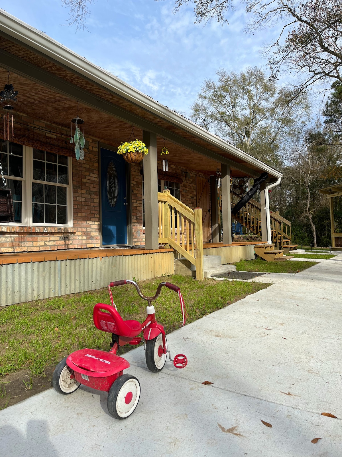 A red tricycle rests on a concrete driveway, positioned in front of a charming brick cottage. The welcoming entrance is accentuated by a blue door and hanging plants. Wooden steps lead up to a spacious porch, surrounded by lush green grass.