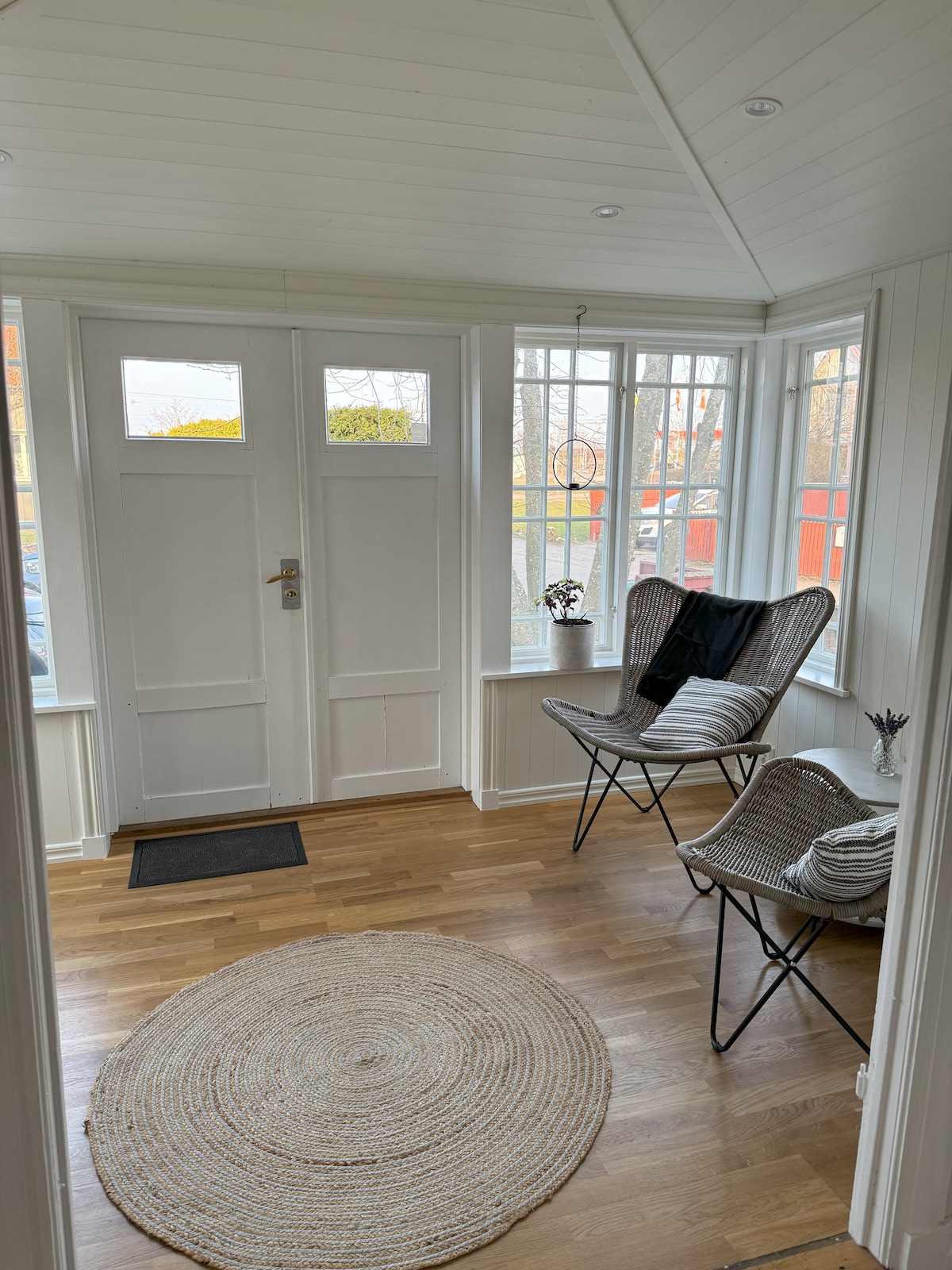 A bright entryway features two woven chairs positioned beside large windows, allowing natural light to fill the space. A round, textured rug lies on the wooden floor, adding warmth. The entry is enhanced by simple decor and a welcoming atmosphere.