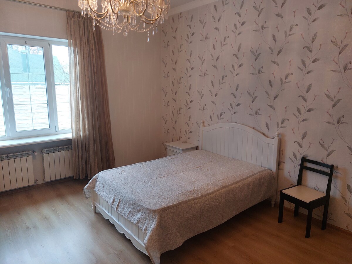A serene bedroom is highlighted by soft wallpaper featuring a floral pattern. A spacious bed is topped with a light-colored coverlet. Natural light filters through a large window, while an elegant chandelier hangs from the ceiling. A small chair is positioned near the bed.
