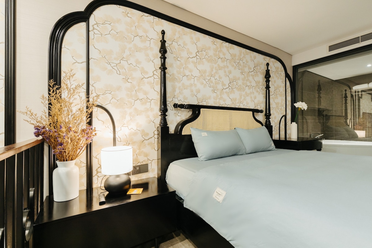 The bedroom features a spacious bed with a soft blue coverlet and a decorative headboard. A bedside table holds a modern lamp, and a vase with flowers adds a touch of nature. Large windows provide natural light, enhancing the calming atmosphere.