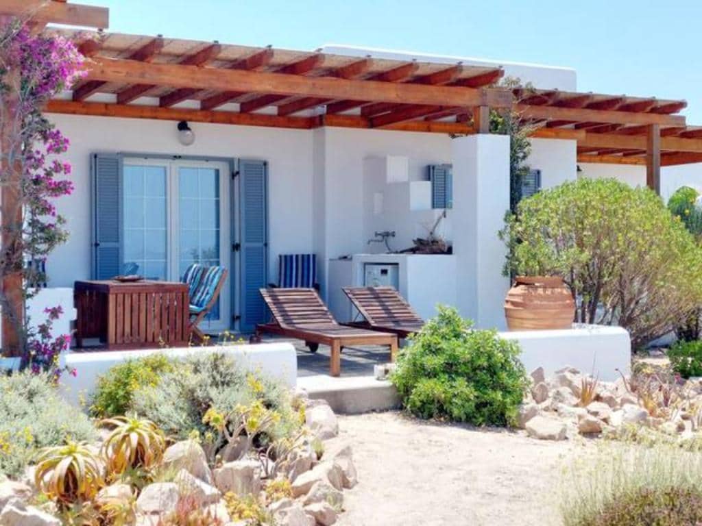 A serene outdoor seating area features two wooden lounge chairs and a small table under a wooden pergola. The space is surrounded by a garden with various plants and stone pathways, and a kitchenette is visible in the background.