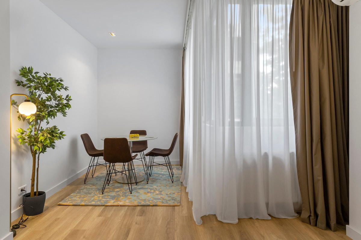 A dining area features a round table surrounded by four brown chairs. Natural light filters through sheer curtains, creating a bright atmosphere. A small potted plant adds a touch of greenery, and a patterned rug lies underneath the table, enhancing the space's warmth.