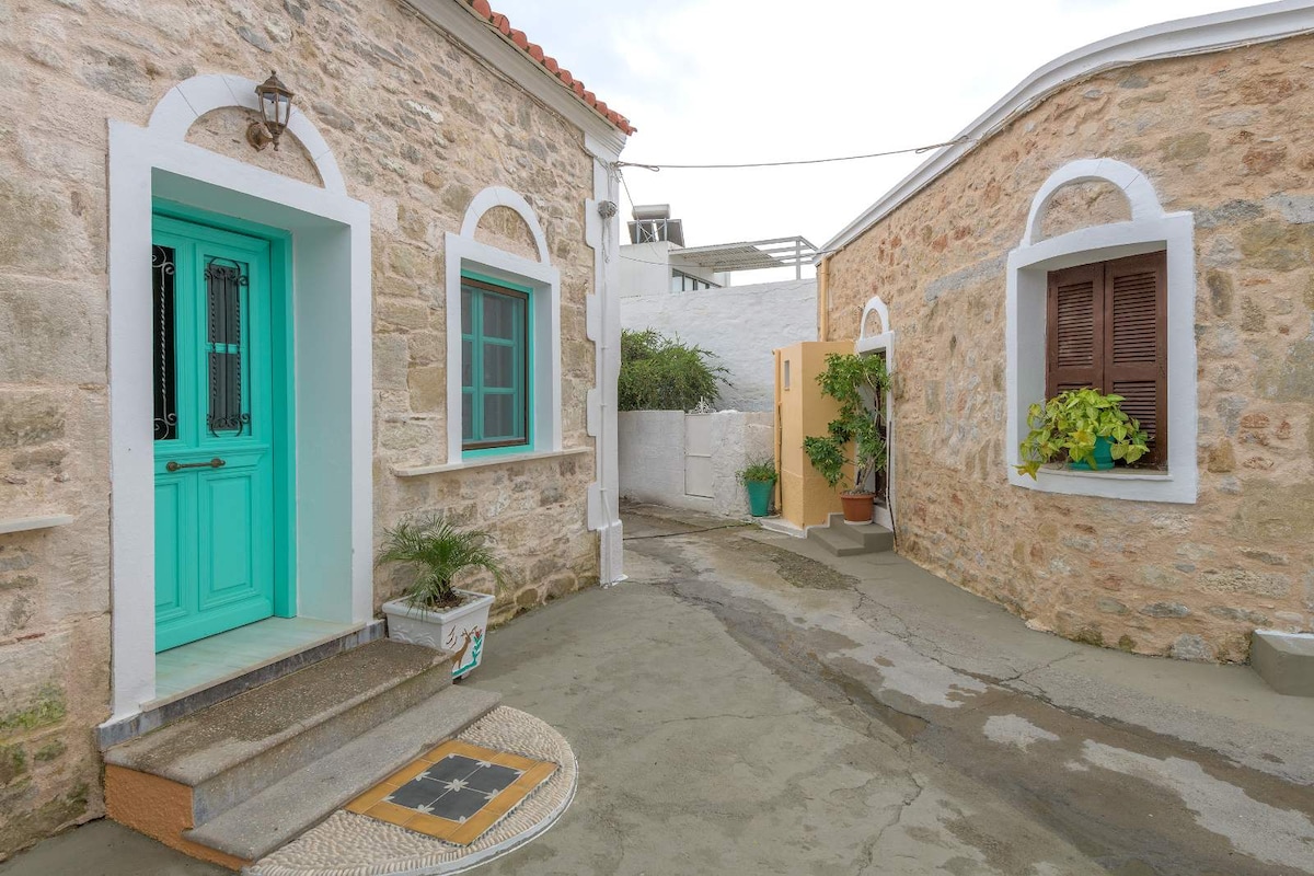 A charming alleyway is lined with traditional stone buildings, showcasing a mint green door and a wooden window framed by white accents. Potted plants add a touch of greenery, while the pathway is paved with a decorative design. The peaceful atmosphere is enhanced by the soft evening light.