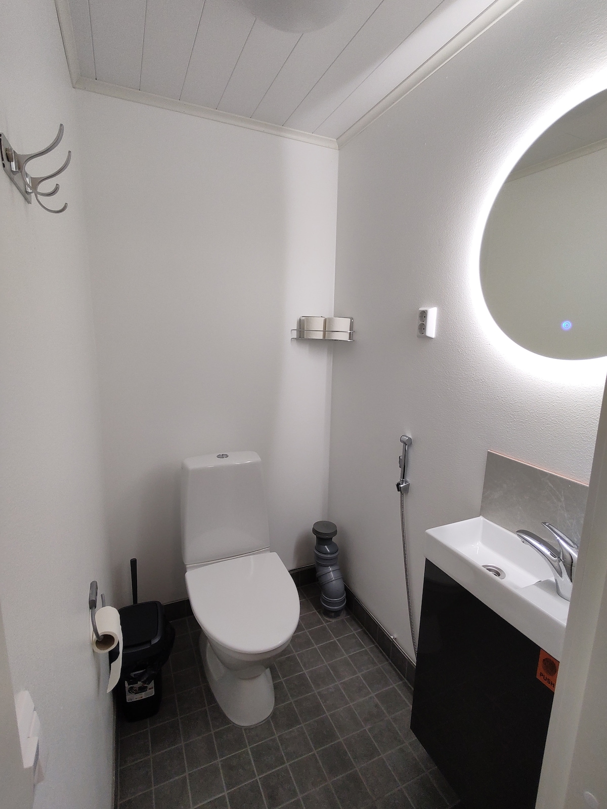 A clean and modern bathroom is presented, featuring a toilet and a small sink with a sleek faucet. A circular mirror above the sink reflects soft light. The dark gray tiled floor contrasts with the bright walls, creating a simple yet functional space.