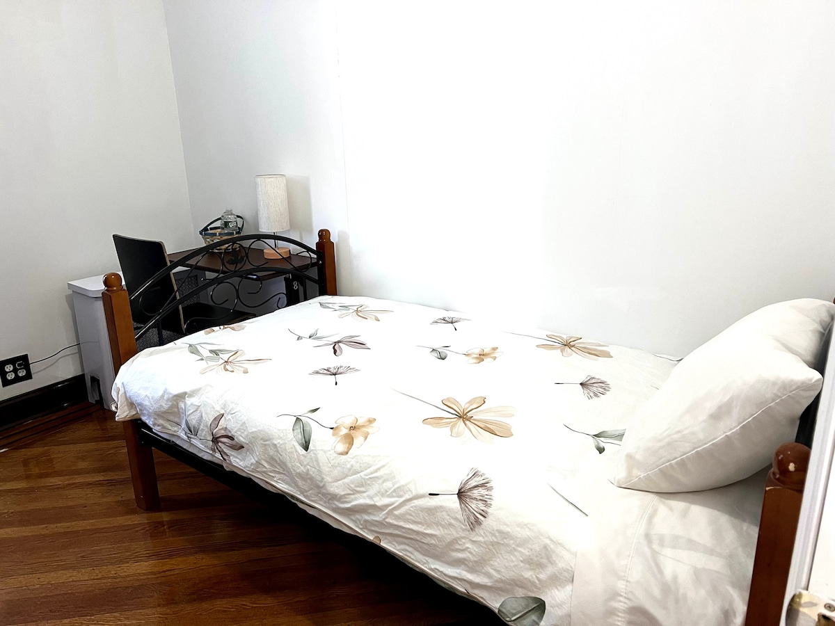 A cozy private room features a twin-sized bed dressed with light-colored bedding adorned with floral patterns. A small desk and lamp are positioned nearby, along with a waste bin. Natural light brightens the space through a nearby window, and the hardwood floor adds warmth.