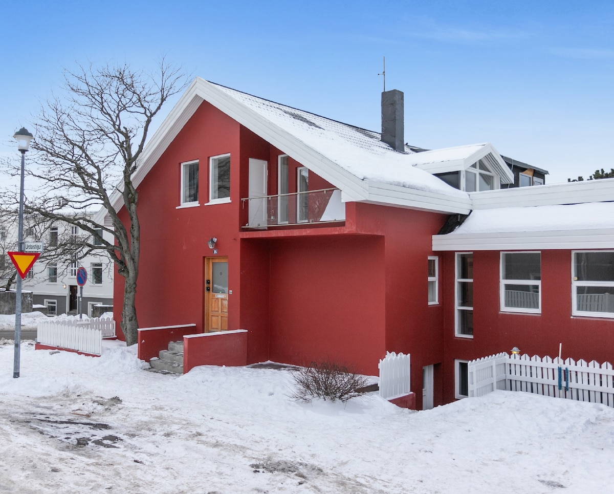 A modern red house is featured with a sloped roof and an angled balcony. White trim accents the windows, and a white picket fence surrounds the property. Snow covers the ground, creating a serene winter landscape outside.