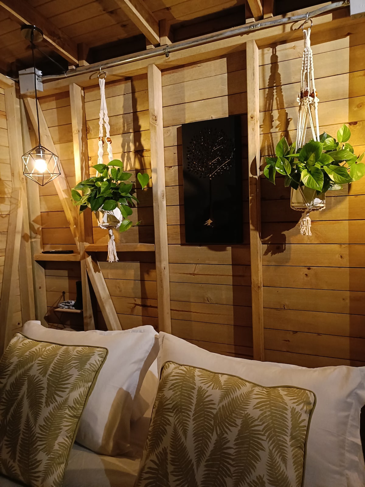 A bedroom corner features a wooden wall adorned with hanging planters filled with greenery. A geometric light fixture illuminates the space, while two decorative pillows with leaf patterns rest on a spacious bed, providing a serene and natural ambiance.