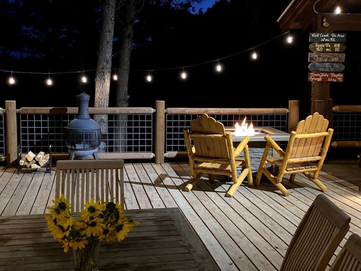 An outdoor deck is illuminated by string lights, set against a dark evening sky. Two wooden Adirondack chairs are positioned around a fire pit, with a blue metal heater nearby. A table features a vase of sunflowers, enhancing the inviting outdoor space.
