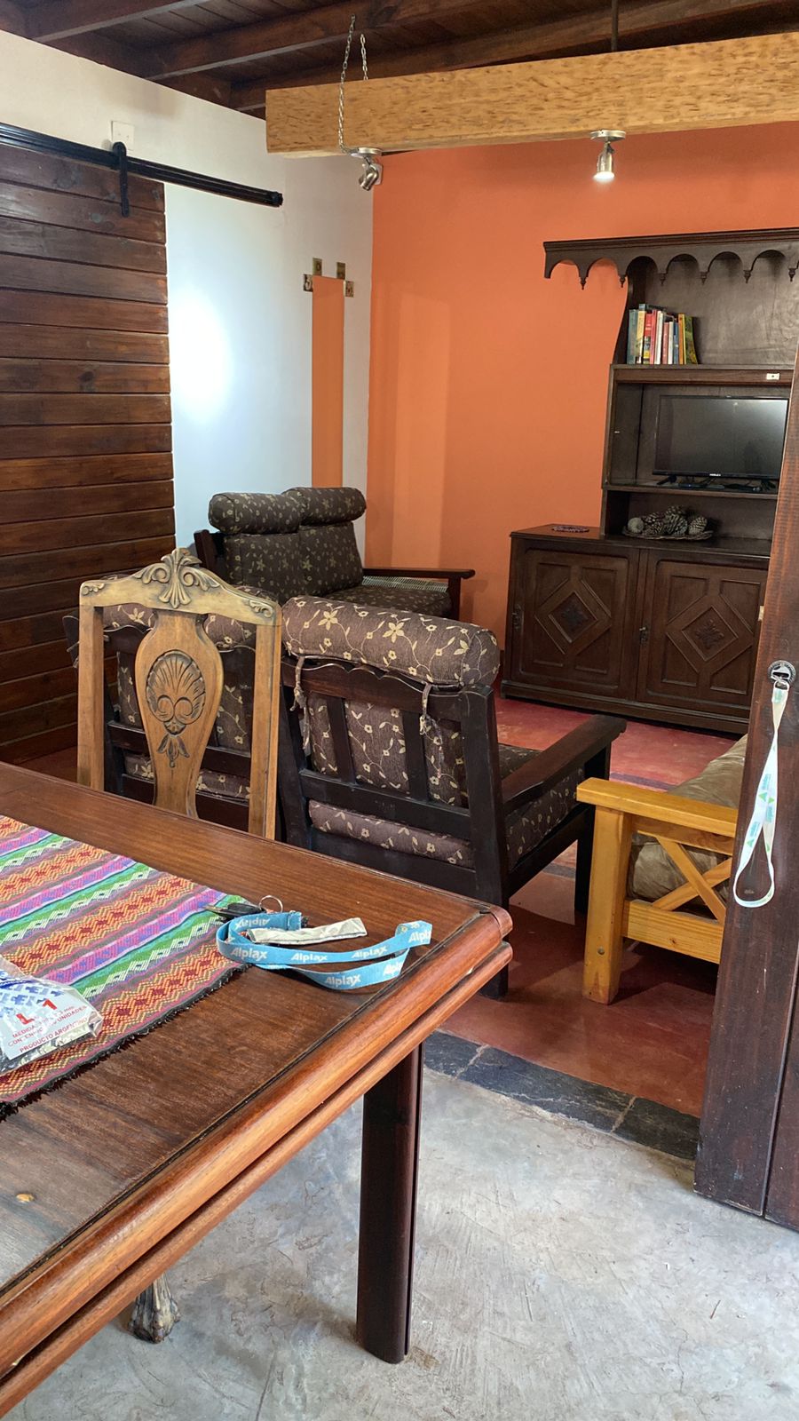 A cozy living area features a wooden table, surrounded by two patterned armchairs and a wooden cabinet. The space is divided by a door, revealing warm orange and dark wood tones that create a welcoming ambiance.