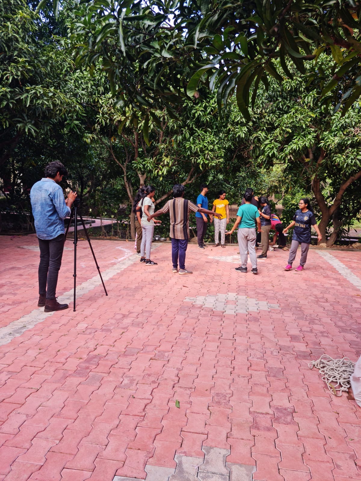 A group of individuals is engaged in a dynamic activity on a pink-paved outdoor area, surrounded by lush greenery. A person is seen photographing the gathering with a camera mounted on a tripod. The atmosphere appears lively and collaborative.