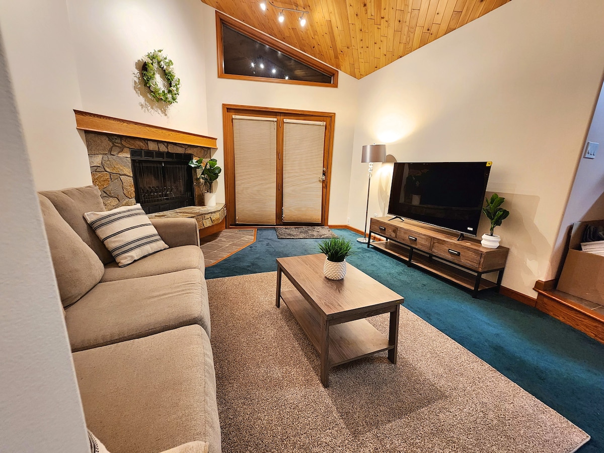 A cozy living area features a comfortable sofa and a wooden coffee table, adorned with a small plant. A stone fireplace serves as a focal point, while large windows invite natural light. A television is positioned on a media console across from the seating.