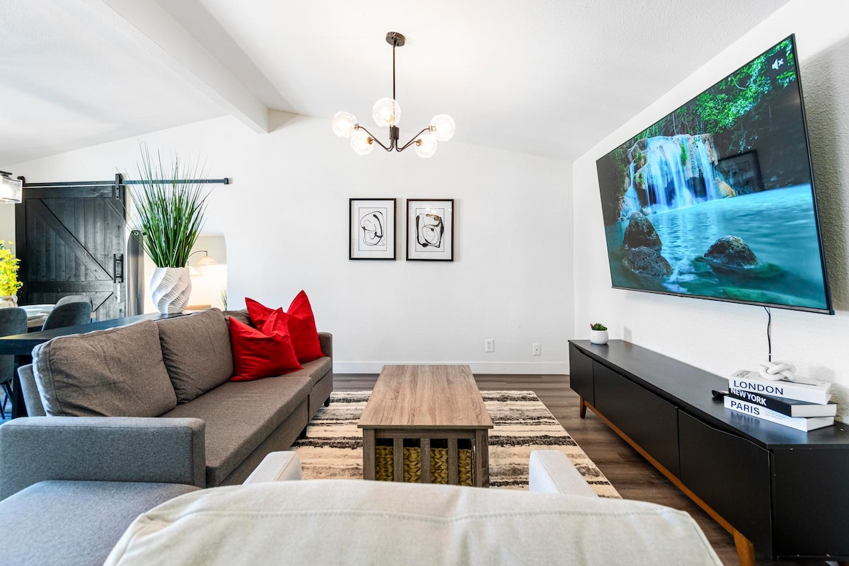 A spacious living area is depicted, featuring a modern grey sectional sofa adorned with red cushions. A large flat-screen TV displays a serene nature scene. A stylish coffee table rests on a patterned rug, complemented by a floor lamp and decorative plants in the background.