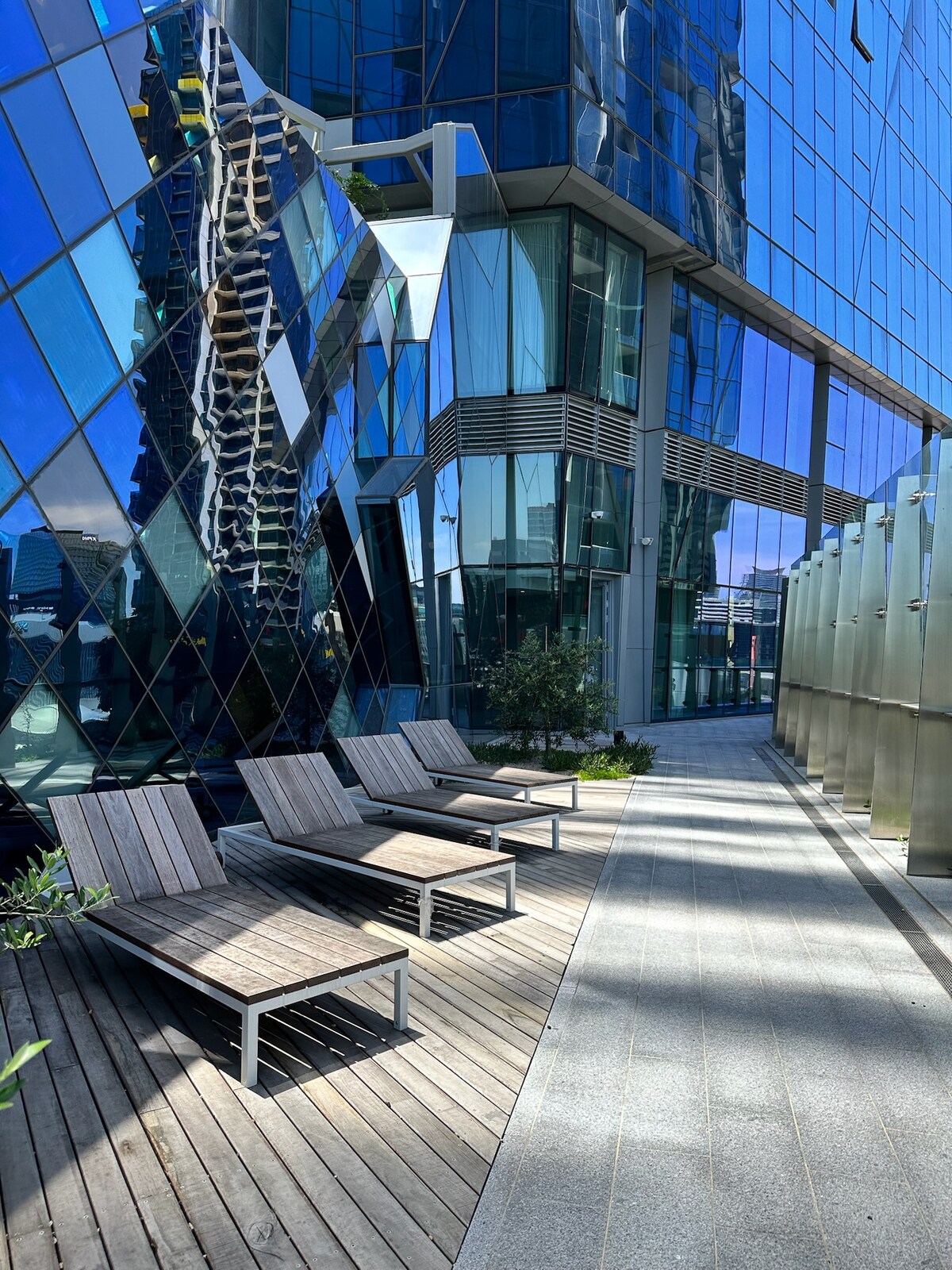 Lounge chairs are arranged on a wooden deck, positioned beside a sleek glass facade. The exterior reflects the surrounding skyline and cityscape, with sunlight illuminating the area. Greenery is incorporated into the design, enhancing the outdoor space.