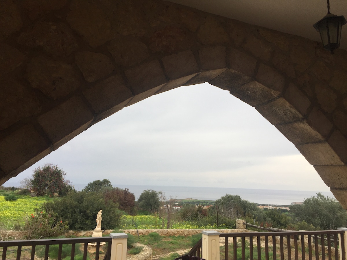 An arched stone entrance frames a view of the serene sea and lush greenery. The foreground features a wooden railing and a sculpture, while vibrant fields extend toward the horizon, meeting the calm waters under a soft, cloudy sky.