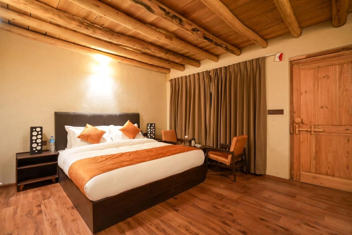 A spacious bedroom features a large bed with an orange and white duvet. Two decorative pillows sit atop the bed. Wooden beams span the ceiling, and natural light filters through the curtains. A small table with two chairs is positioned near the window.