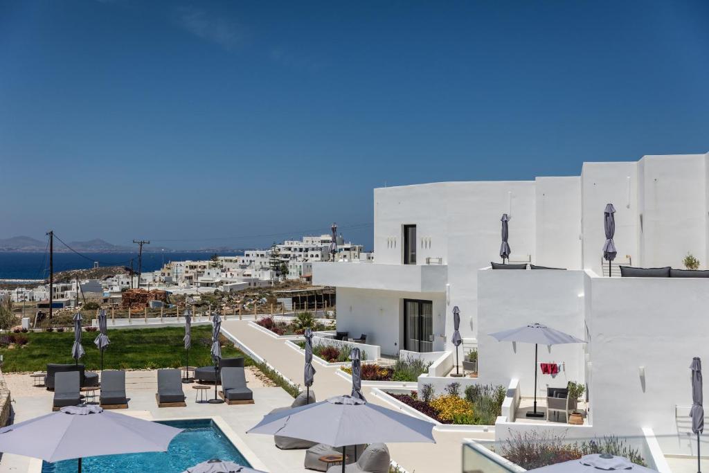 A serene outdoor area features a shared pool surrounded by lounge chairs and umbrellas. The white building contrasts with the vibrant greenery and the blue sea in the background. Clear skies enhance the bright and open atmosphere of the space.
