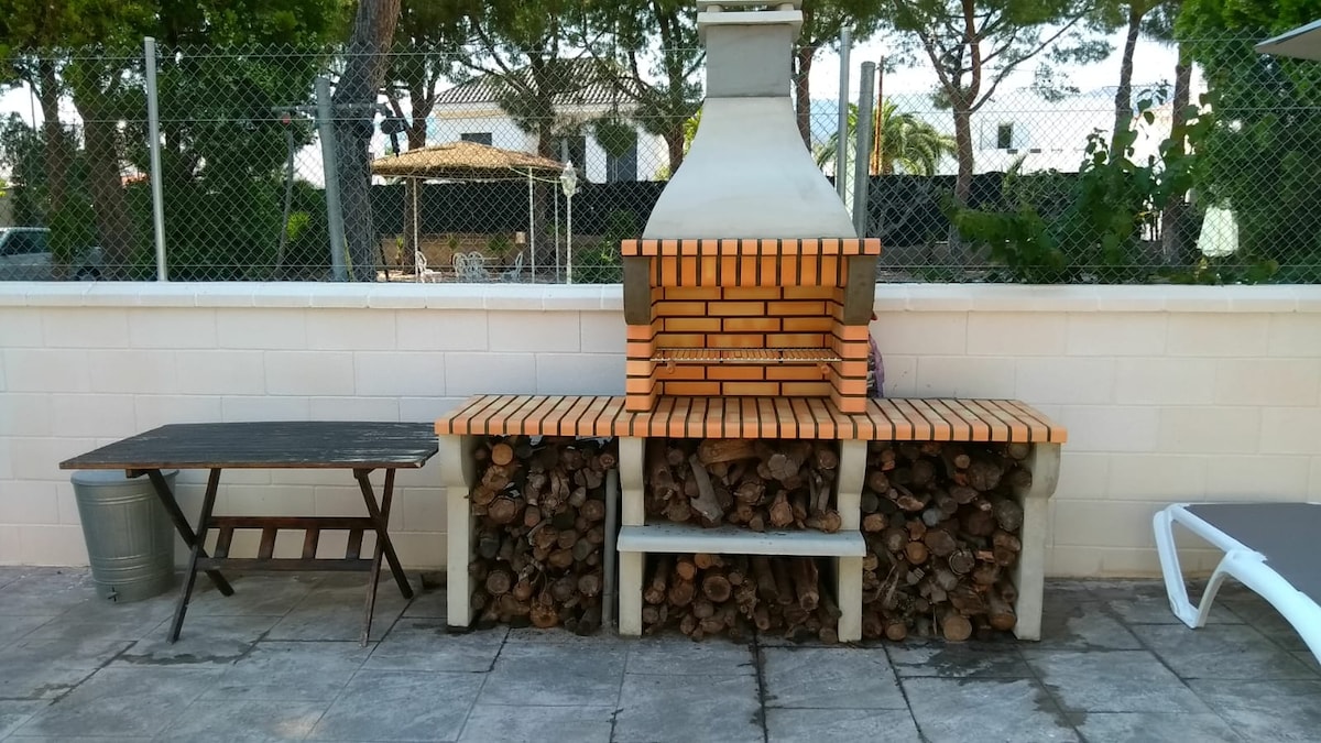 An outdoor barbecue area is presented with a brick grill, complemented by a wooden storage space for logs. A metal container is placed nearby, and a folding table is positioned to the side. Sunlight filters through the surrounding trees, enhancing the relaxed setting.