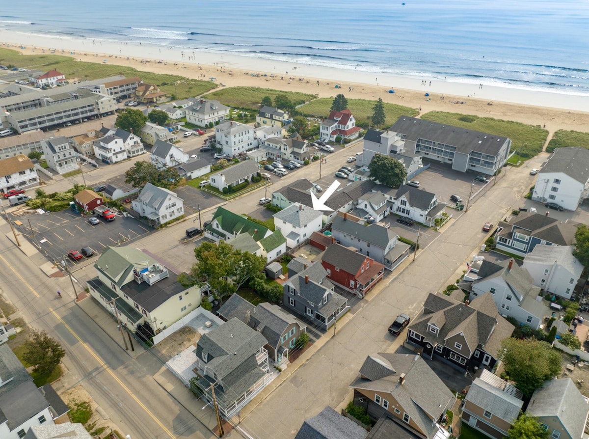 An aerial view captures the cottage's prime location just steps from the sandy beach. Nearby structures and the expansive shoreline are visible, showcasing the area’s vibrant seaside atmosphere. The image illustrates the proximity to both the beach and surrounding establishments.