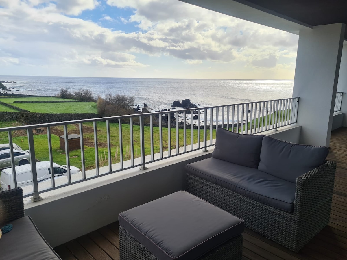 A covered balcony features comfortable outdoor seating consisting of a plush sofa and an ottoman. The view encompasses the ocean and lush green landscape, with the horizon showcasing a partly cloudy sky.