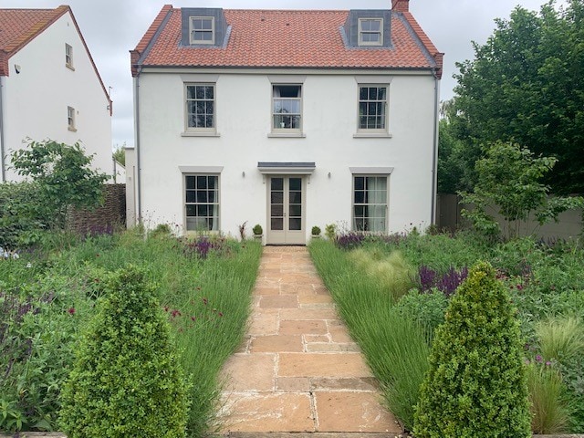 A two-story house is set against a green backdrop, featuring a central entrance adorned with a pair of potted plants. A stone pathway leads through a lush garden filled with various plants and flowers, enhancing the home's natural appeal.