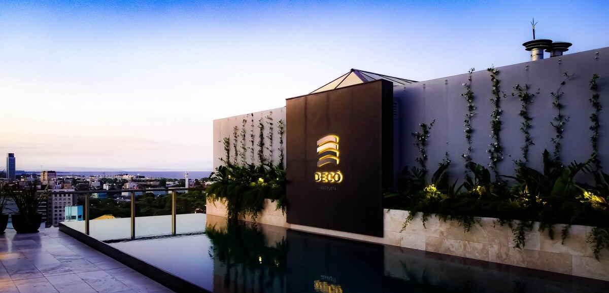 A rooftop area features a reflective pool bordered by lush greenery and modern landscaping. A prominent sign displays the property name, set against a serene evening sky transitioning from light to twilight. The surrounding cityscape is visible in the distance.