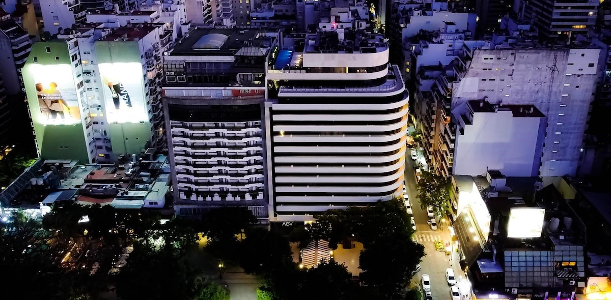 An aerial view captures the modern architecture of a prominent building, showcasing curved lines and illuminated windows. Surrounding structures contribute to the vibrant urban atmosphere, while city streets are lined with vehicles and trees, emphasizing the bustling environment of the neighborhood.
