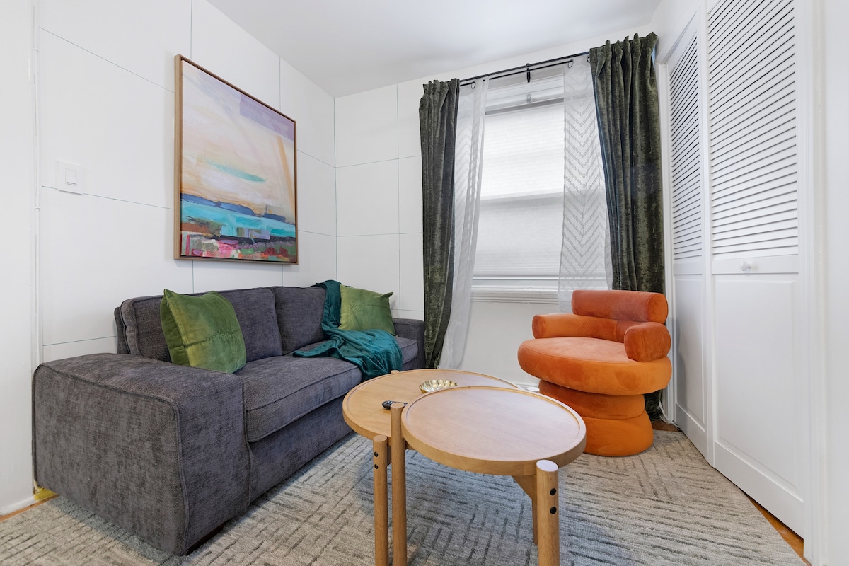A cozy seating area is presented with a dark grey sofa and a vibrant orange chair, complemented by a round wooden coffee table. Natural light filters through sheer curtains, highlighting the artistic wall painting and textured rug beneath.