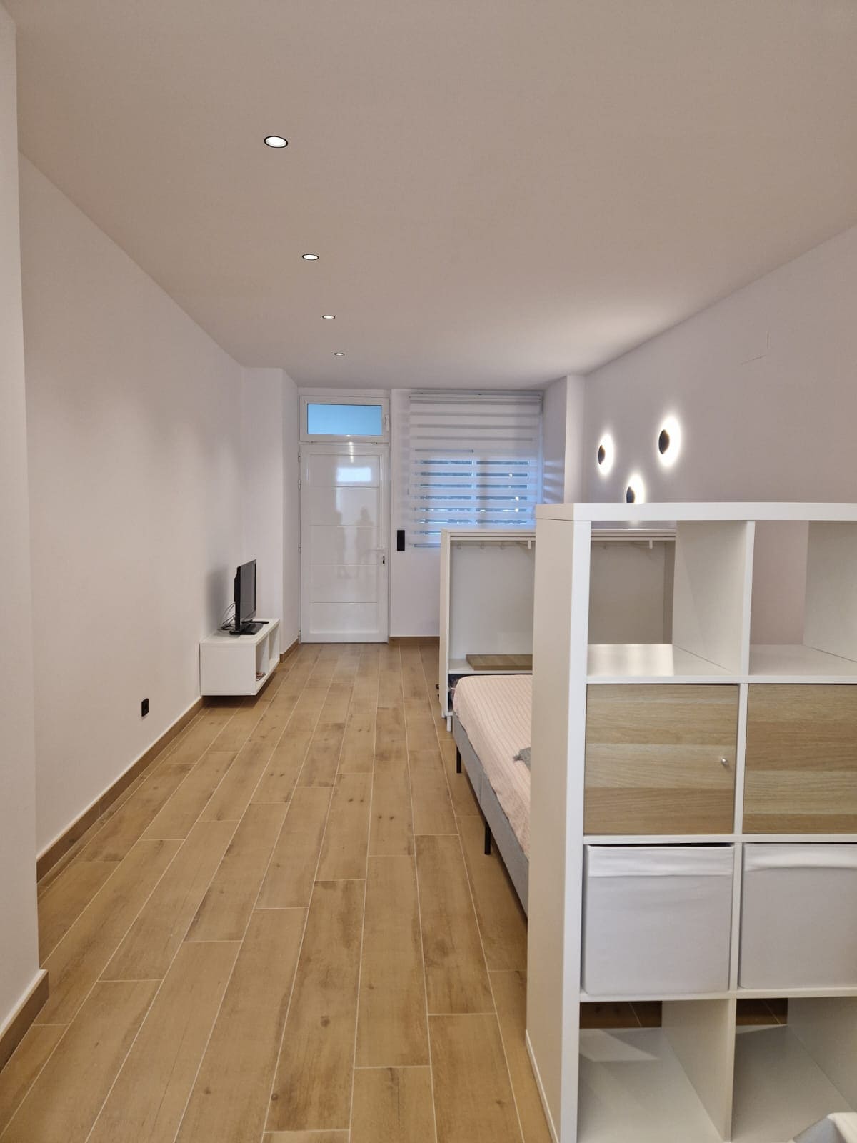 A spacious room is presented with light wood flooring and soft white walls. A comfortable double bed is separated by a storage unit from the living area, featuring a flat-screen television. Natural light enters through a window adorned with blinds.