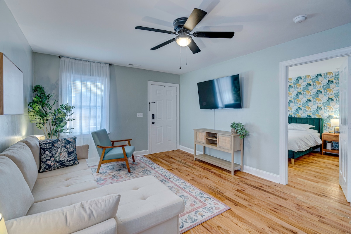 A bright living area features a light-colored sofa and an accent chair, set against pale green walls. A ceiling fan provides airflow, while natural light enters through a window adorned with sheer curtains. A television is mounted on the wall opposite the seating arrangement.
