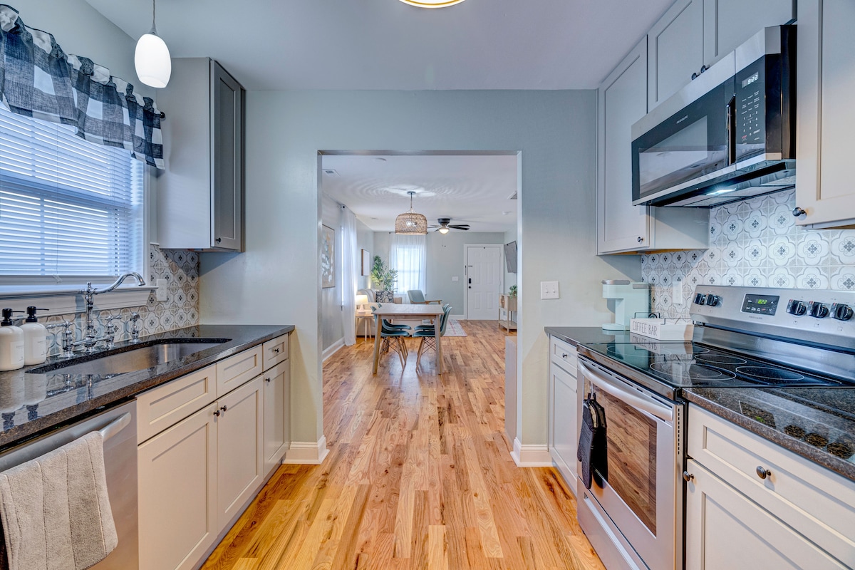 A modern kitchen is presented with light cabinetry and dark countertops. Stainless steel appliances are visible, including an oven and microwave. The dining area can be seen in the background, featuring a round pendant light and a table with seating. Natural light filters through the window.