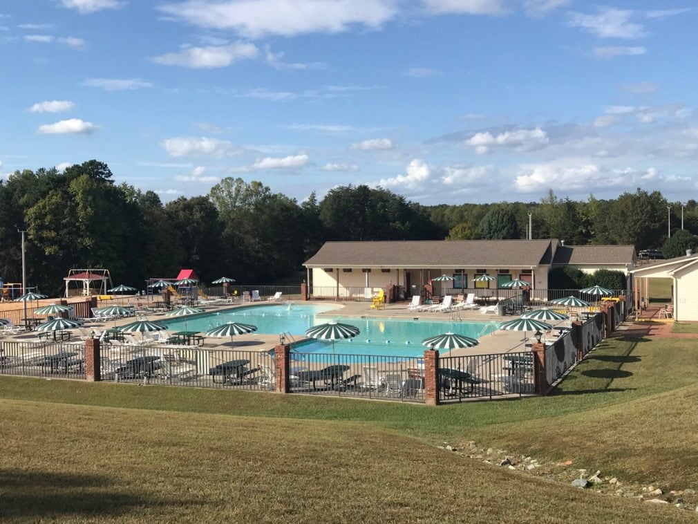 An expansive view of the Olympic-sized saltwater pool is showcased, surrounded by lounge chairs and green-and-white striped umbrellas. A nearby building provides access to amenities, with grassy areas visible in the background, under a partly cloudy sky.