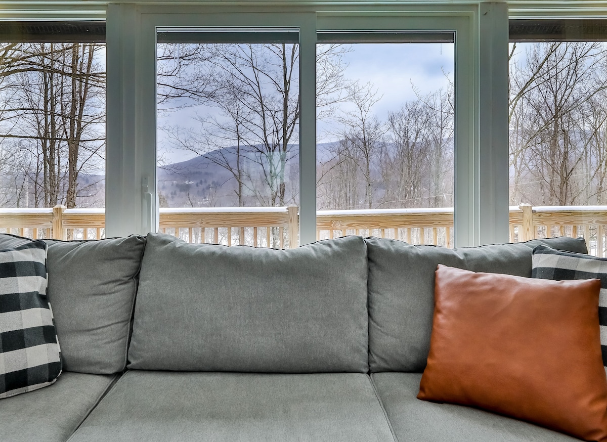 A spacious gray couch is positioned in front of large windows, revealing views of the surrounding trees and distant mountains. A single brown throw pillow adds a touch of warmth to the seating area, inviting relaxation and connection with the outdoor landscape.