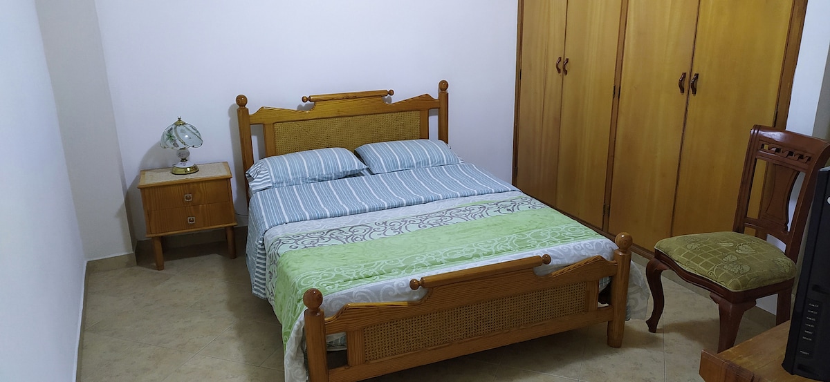 A well-arranged bedroom is featured, showcasing a spacious bed with striped bedding and a decorative throw. A bedside table with a lamp stands next to the bed. A wooden wardrobe occupies one wall, complemented by a chair and a small television in the corner.