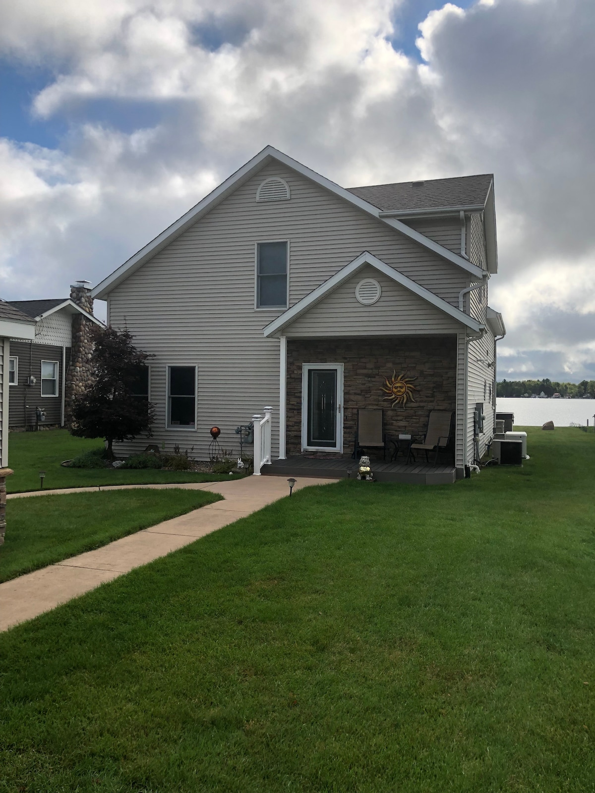A two-story lakefront home is shown, featuring a light-colored exterior with stone accents. A welcoming pathway leads to the front entry. Two chairs are positioned on the patio, with a scenic view of the lake visible beyond the green lawn.