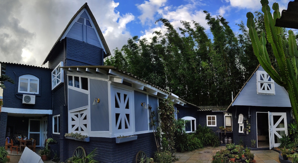 The image displays a charming blue and white house with unique architectural features, surrounded by lush greenery. Bamboo trees sway gently in the background, while a spacious courtyard with stone flooring and various potted plants is visible, providing a serene outdoor setting.