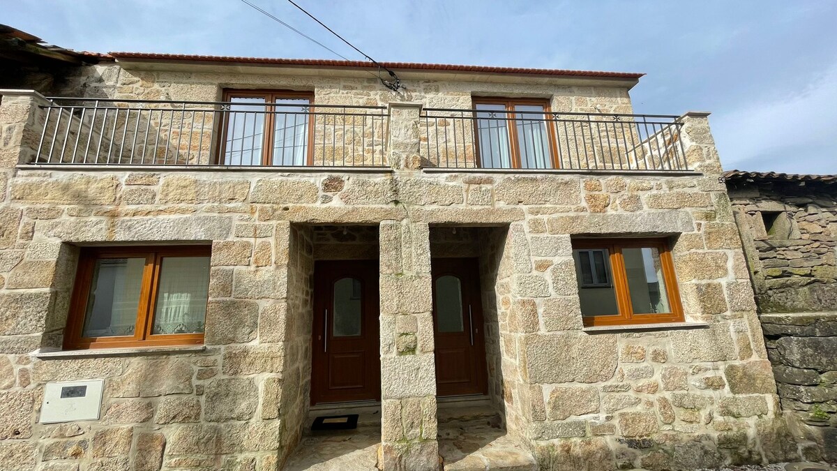 A charming stone exterior of a two-story house features a symmetrical facade with wooden doors and large windows. A balcony spans the upper level, providing an open view. The natural stonework enhances the rustic character of the property, rooted in the local countryside.