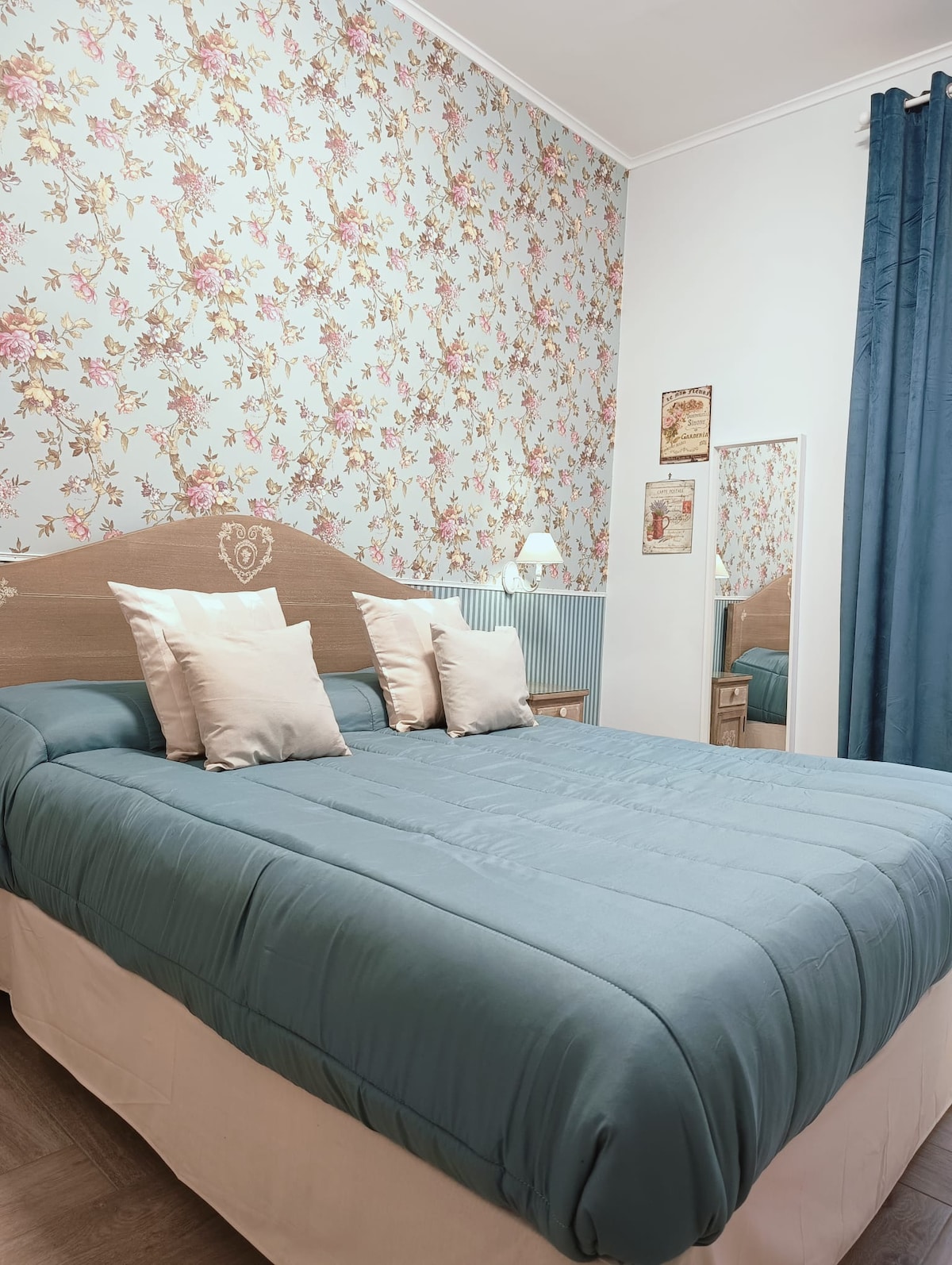 A bedroom features floral wallpaper in soft hues, showcasing a spacious bed with a light blue quilt and decorative pillows. A full-length mirror and bedside lamp add functionality, while dark curtains frame a window, contributing to the room's tranquil ambiance.