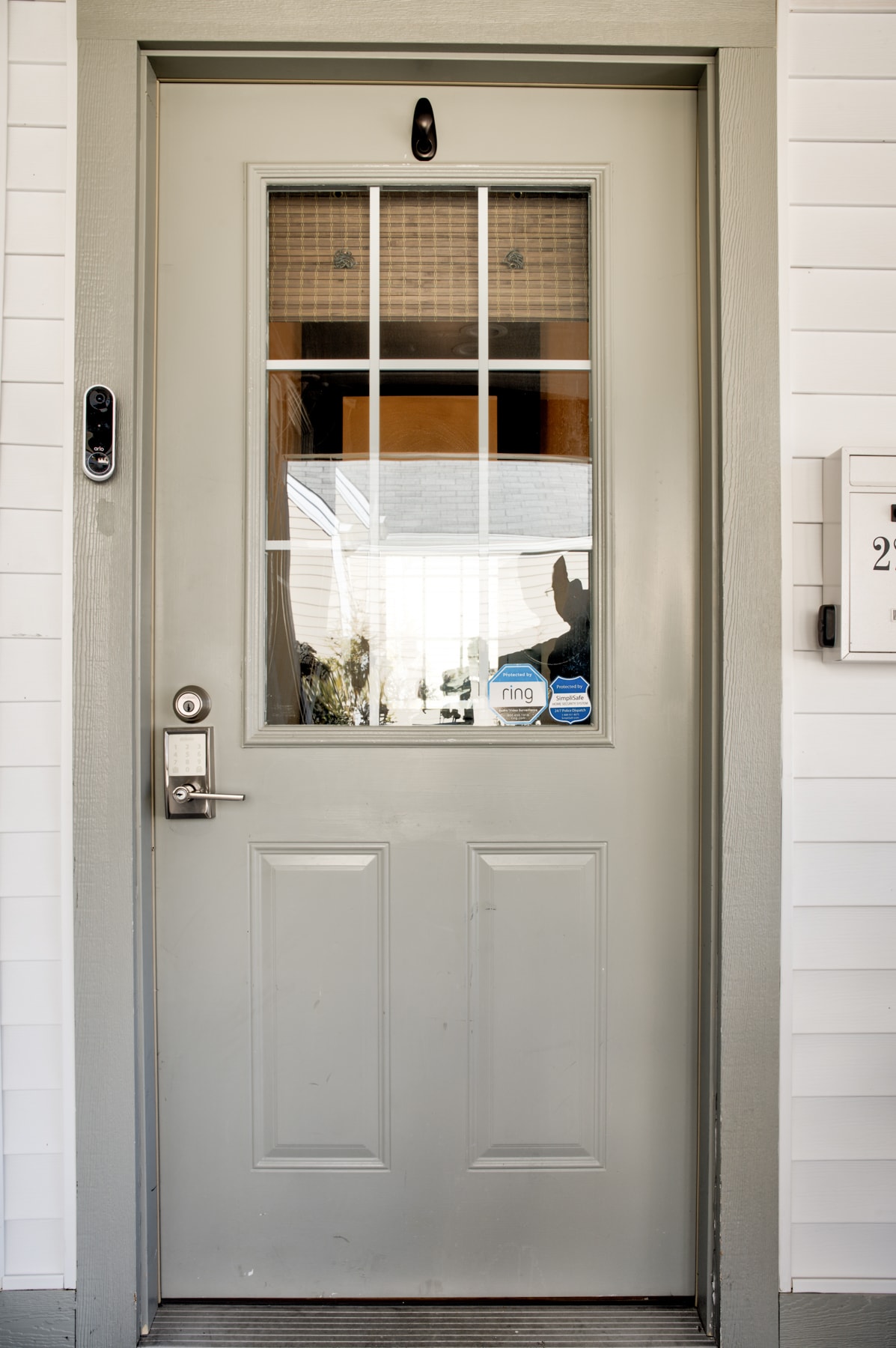 A sturdy light gray door stands as the entrance to the home, featuring four glass panes at the top. A small security camera is mounted on the left side, and a doorbell is positioned below it. A modern mailbox is affixed to the right.