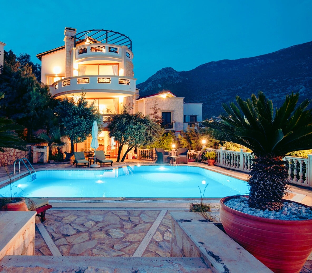 The villa is illuminated at dusk, showcasing a private pool surrounded by lush gardens and stone pathways. The multi-level structure features a circular design with a spacious terrace. Mountains rise in the background, creating a serene environment for relaxation.