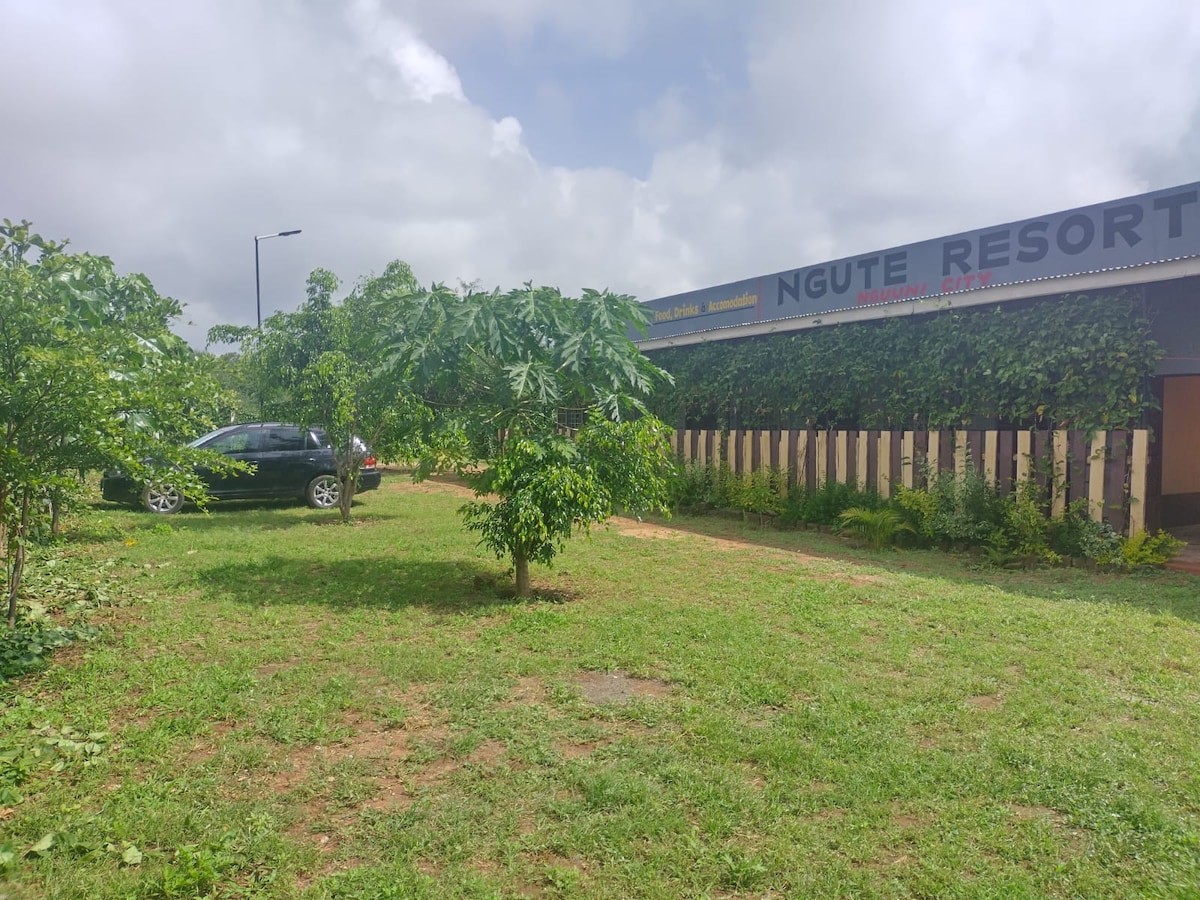A grassy area surrounds Ngute Resort, featuring small trees and shrubs that enhance the natural setting. A dark vehicle is parked near the entrance, which is partially obscured by lush greenery. The building is adorned with climbing plants, blending with the tranquil landscape.
