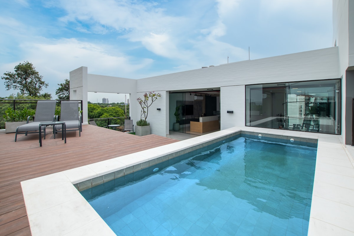 An outdoor area features a refreshing pool surrounded by a sleek wooden deck. Comfortable lounge chairs are positioned nearby, overlooking lush greenery and a modern interior through large glass doors. The space is bathed in natural light under a clear sky.