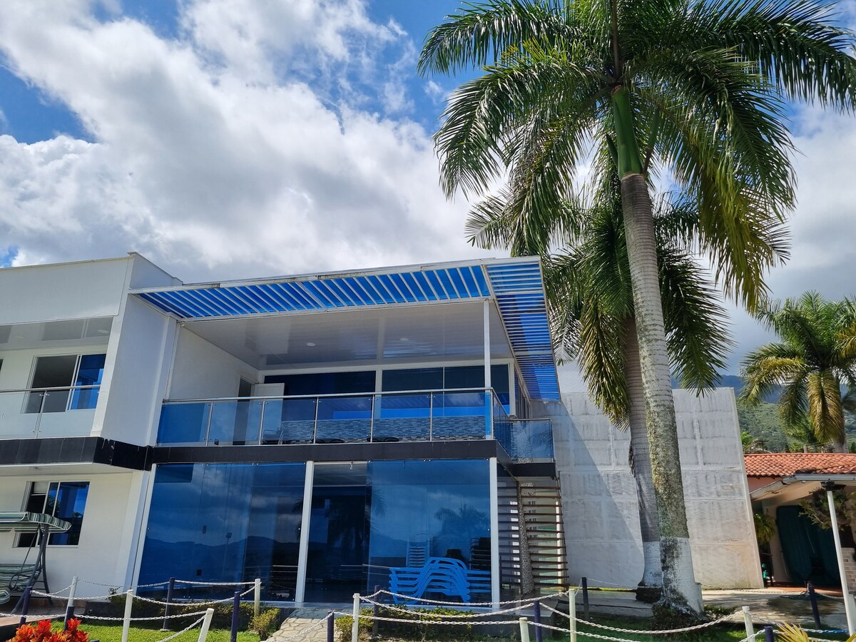 A modern two-story building is showcased, featuring a spacious balcony with blue awnings. Lush palm trees stand nearby, adding to the tropical atmosphere. The exterior reflects large glass windows, allowing for natural light and views of the surrounding area.