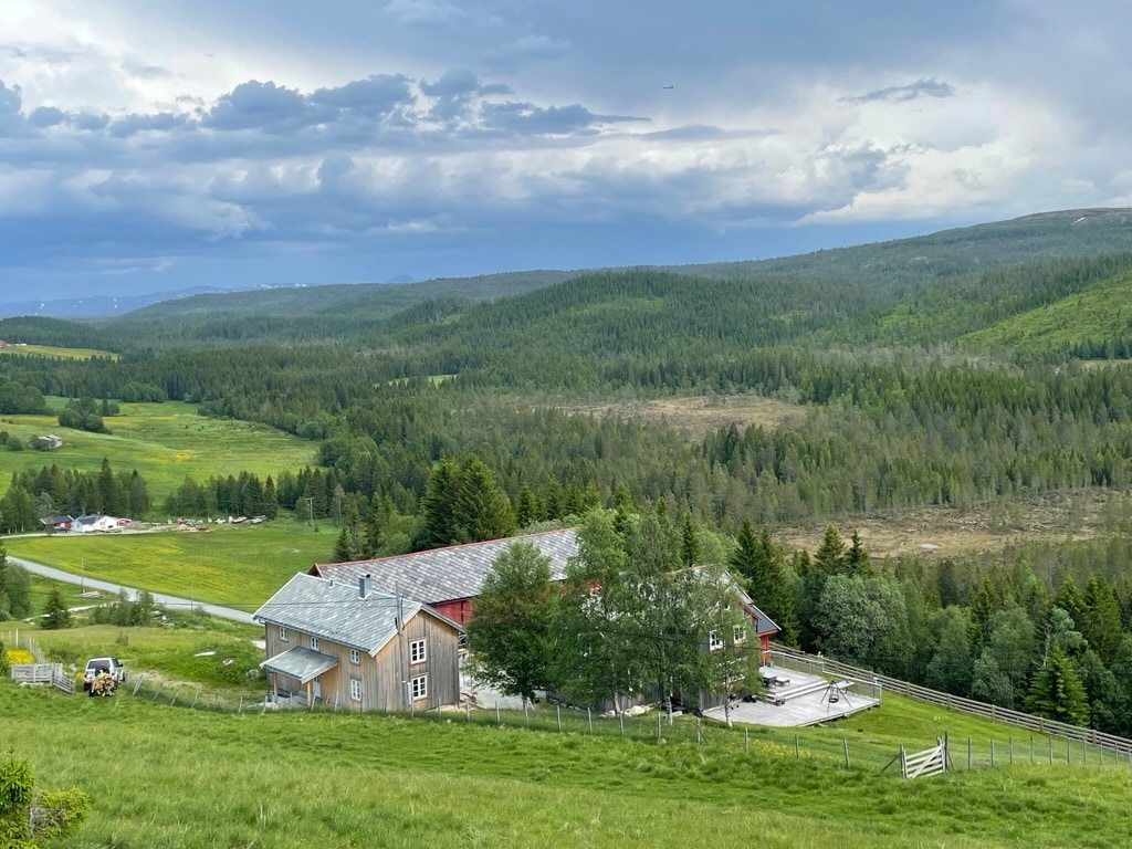 A view of a house set in a lush, green landscape surrounded by rolling hills and dense forests. The structure features a traditional design with a red roof and large outdoor seating areas. The expanse of nature provides a serene backdrop, enhancing the sense of tranquility.